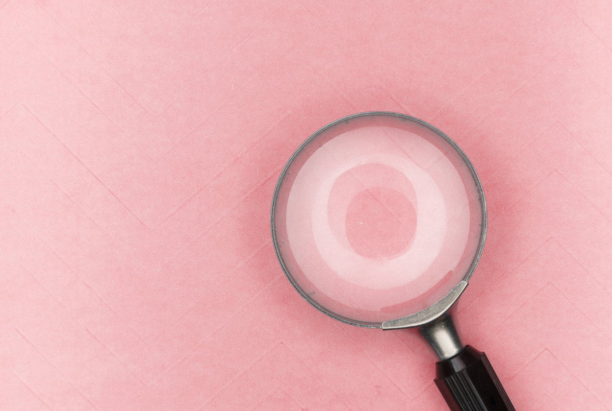 Une loupe posée sur un fond rose poudré : pour savoir si l'on a de la corne, un durillon, un cor, un œil-de-perdrix ou une verrue plantaire, il faut regarder de près la forme, l'endroit et les petits points noirs.