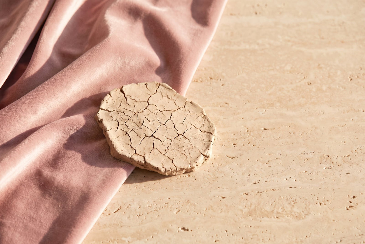 Un morceau d'argile sèche et craquelée posé sur un velours rose poudré : le contraste entre une peau dure qui se fend et une peau souple, comme la corne sur les pieds