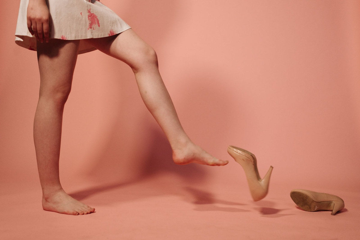 Un pied nu qui vient d'envoyer valser son escarpin beige sur un fond rose : les talons hauts et les chaussures serrées augmentent la pression sous l'avant du pied et favorisent la métatarsalgie et le névrome de Morton.