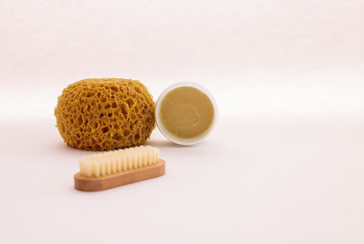 Une éponge naturelle, une petite brosse en bois et un pot de baume ouvert sur un fond rosé : les gestes simples et réguliers de la routine des pieds doux, laver, prendre soin et nourrir la peau.